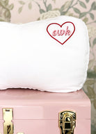 White pillow with 'elk' embroidered in a heart on a pink suitcase with gold accents against a floral background.